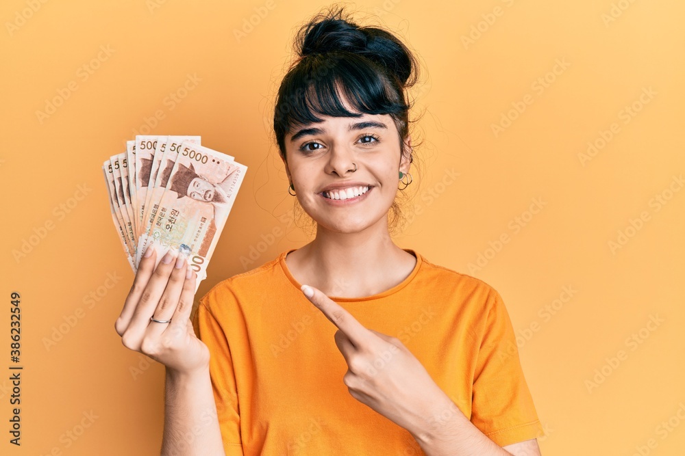 Young hispanic girl holding 5000 south korean won banknotes smiling happy pointing with hand and finger Young hispanic girl holding 5000 south korean won banknotes smiling happy pointing with hand and finger