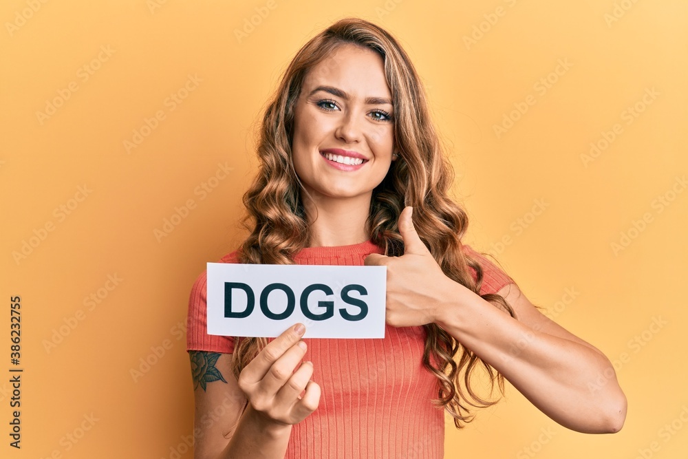 Young blonde girl holding paper with dogs message smiling happy and positive, thumb up doing excellent and approval sign Young blonde girl holding paper with dogs message smiling happy and positive, thumb up doing excellent and approval sign