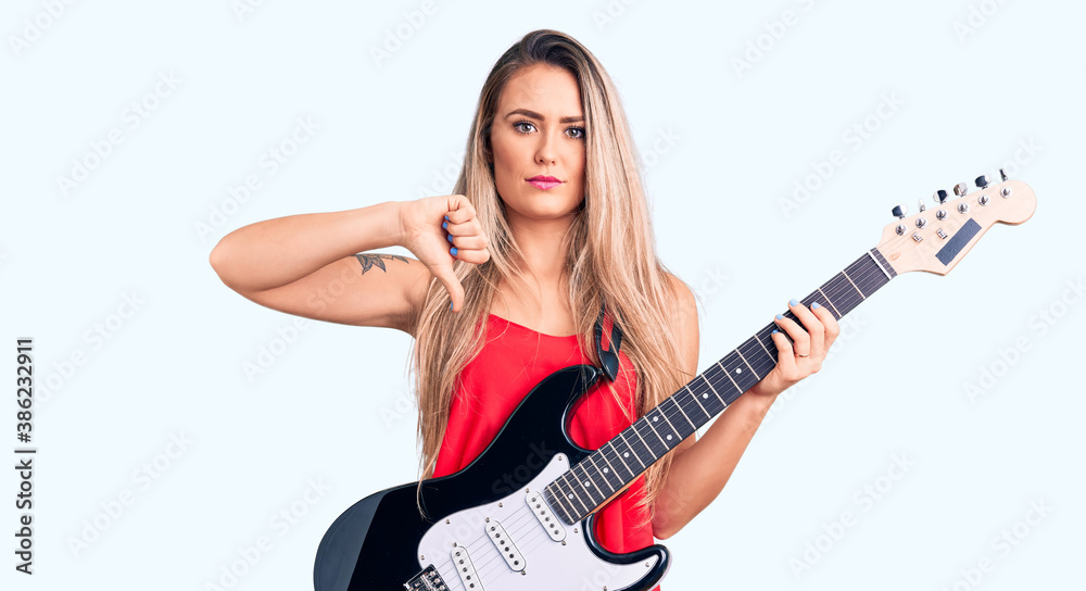 Young beautiful blonde woman playing electric guitar with angry face, negative sign showing dislike with thumbs down, rejection concept