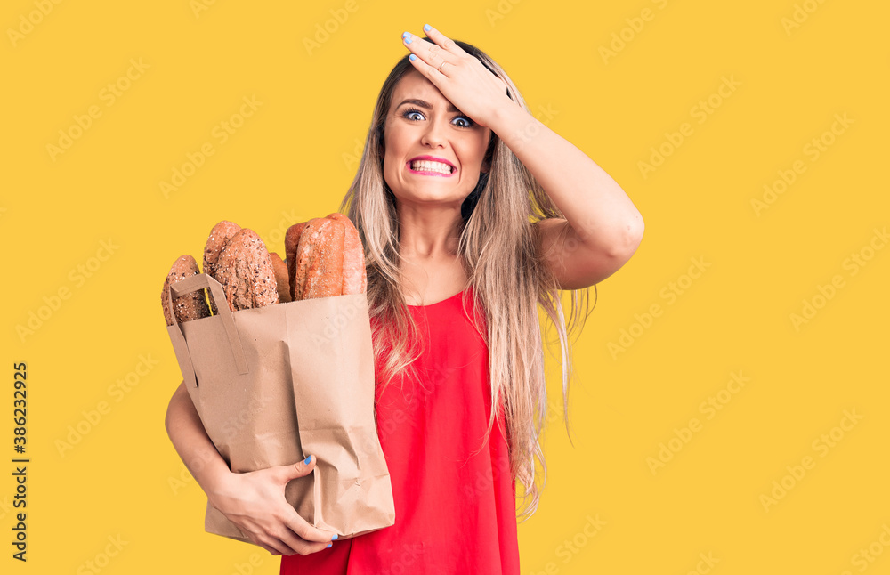 Young beautiful blonde woman holding paper bag with bread stressed and frustrated with hand on head, surprised and angry face
