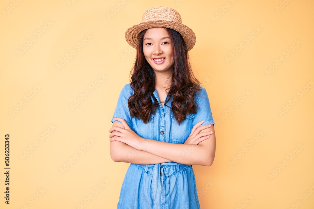 Young beautiful chinese girl wearing summer hat happy face smiling with crossed arms looking at the camera. positive person. Young beautiful chinese girl wearing summer hat happy face smiling with crossed arms looking at the camera. positive person.
