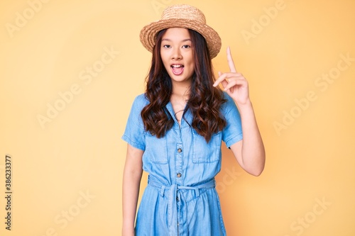Young beautiful chinese girl wearing summer hat pointing finger up with successful idea. exited and happy. number one.