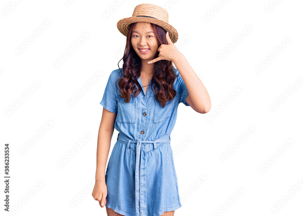 Young beautiful chinese girl wearing summer hat smiling doing phone gesture with hand and fingers like talking on the telephone. communicating concepts. Young beautiful chinese girl wearing summer hat smiling doing phone gesture with hand and fingers like talking on the telephone. communicating concepts.
