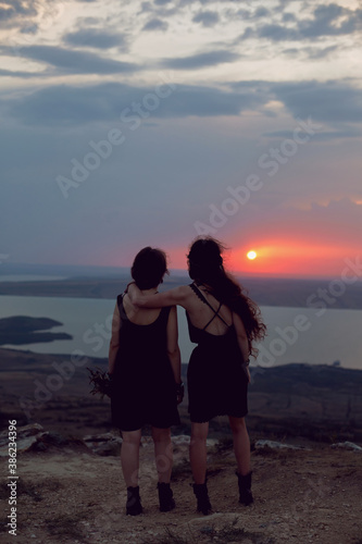 two woman in black dresses walk on the mountain with a bouquet