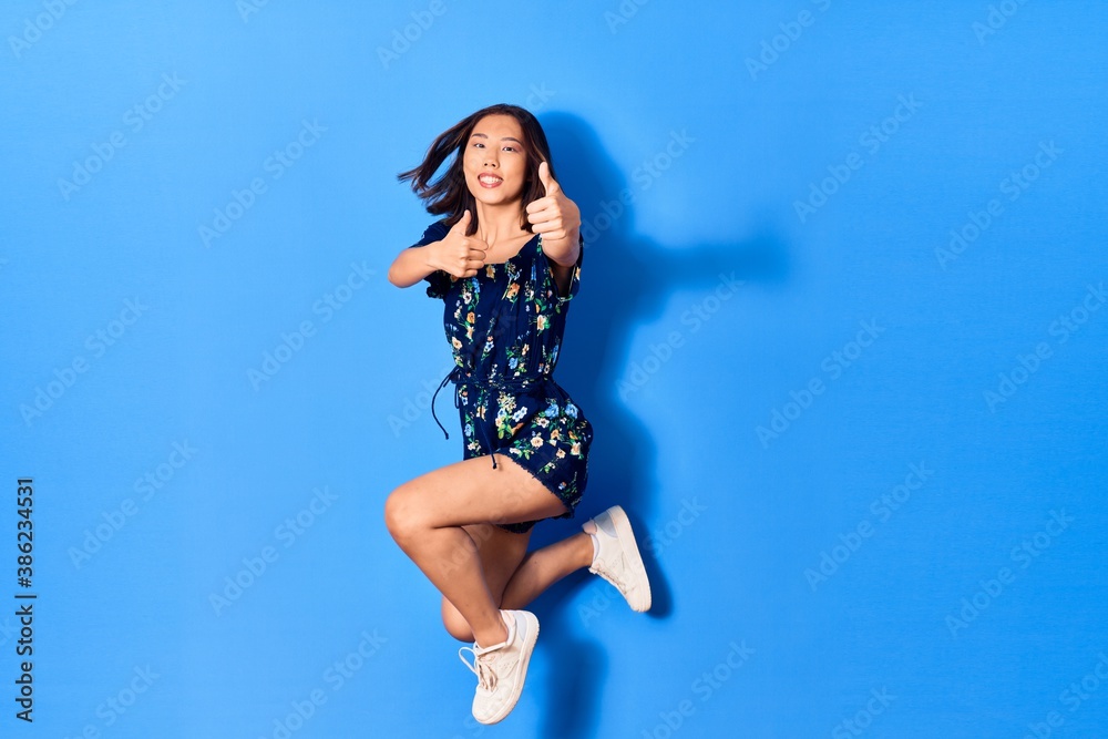 Young beautiful chinese woman wearing casual clothes smiling happy. Jumping with smile on face doing ok sign with thumbs up over isolated blue background Young beautiful chinese woman wearing casual clothes smiling happy. Jumping with smile on face doing ok sign with thumbs up over isolated blue background