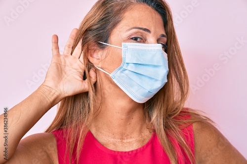 Middle age hispanic woman wearing medical mask smiling with hand over ear listening and hearing to rumor or gossip. deafness concept. photo