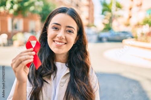 Фотография Young hispanic woman smiling happy holding red hiv ribbon walking at the city