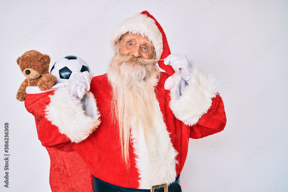 Old senior man with grey hair and long beard wearing santa claus costume holding mustache smiling with a happy and cool smile on face. showing teeth.