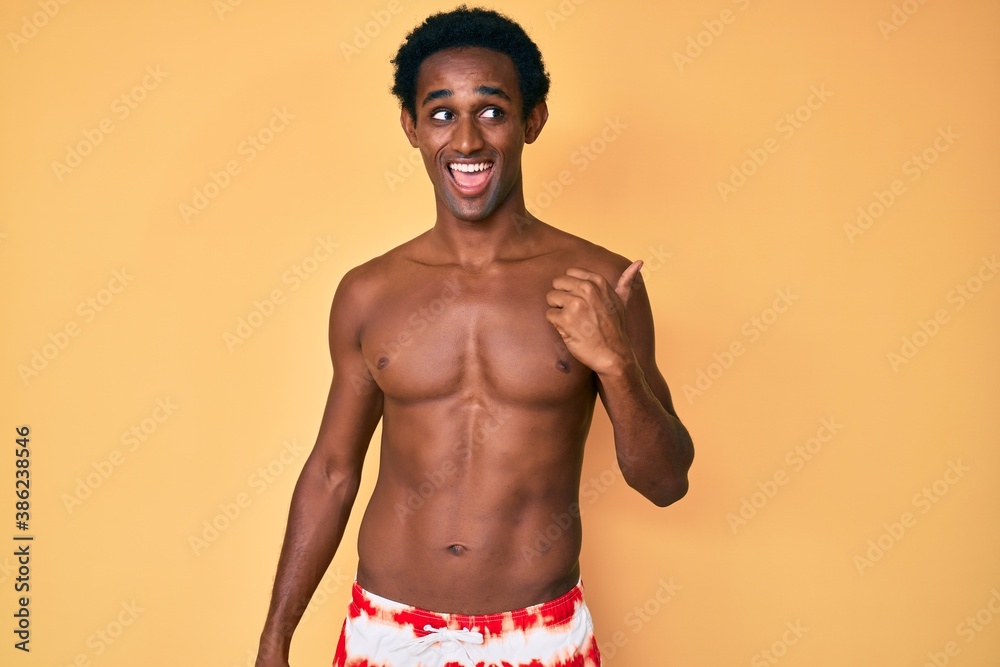 African handsome man wearing swimsuit smiling with happy face looking and pointing to the side with thumb up.