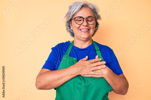 Senior hispanic woman wearing apron and glasses smiling with hands on chest with closed eyes and grateful gesture on face. health concept.