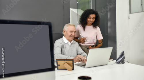 Aged man, senior intern looking at laptop while showing results to his young colleague, Friendly female worker mentoring and training new employee, monitoring his progress at work