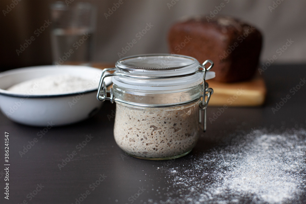 Active rye sourdough starter in a glass jar, a bowl with flour and fresh baked wheat and rye sourdough bread. Active rye sourdough starter in a glass jar, a bowl with flour and fresh baked wheat and rye sourdough bread.