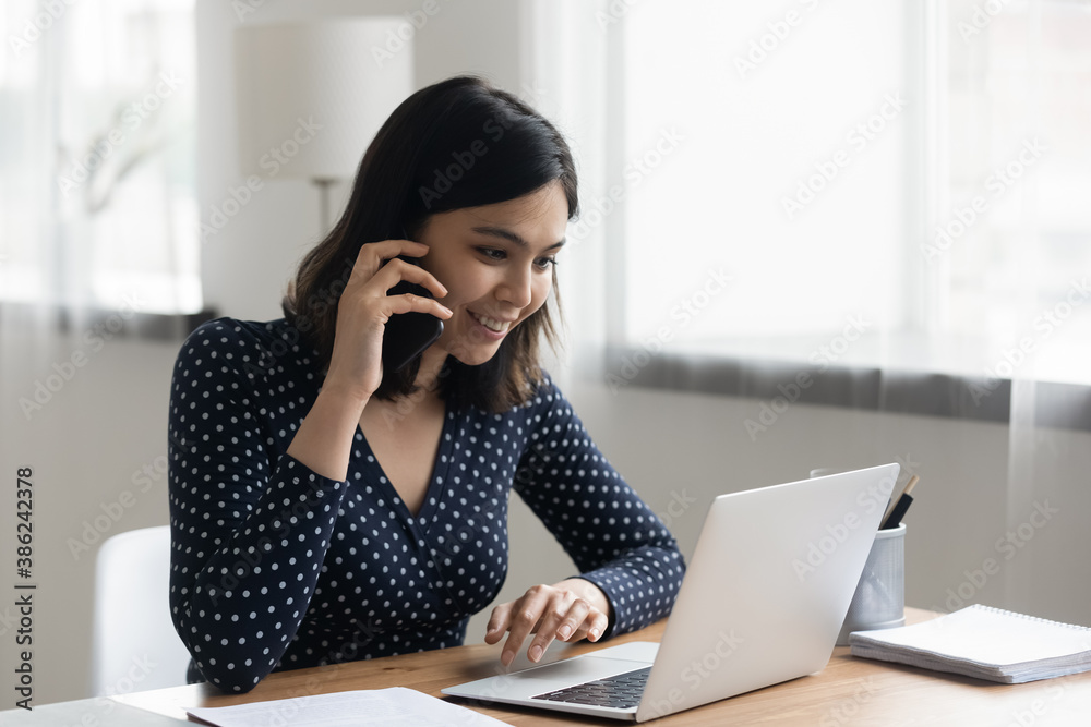 Smiling young asian businesswoman sit at desk at home office look at laptop screen talking on cellphone consulting client online. Happy Vietnamese woman work on computer, have smartphone call. Smiling young asian businesswoman sit at desk at home office look at laptop screen talking on cellphone consulting client online. Happy Vietnamese woman work on computer, have smartphone call.