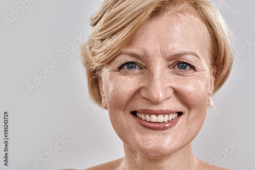 Close up shot of face of beautiful middle aged woman smiling at camera while posing isolated against grey background