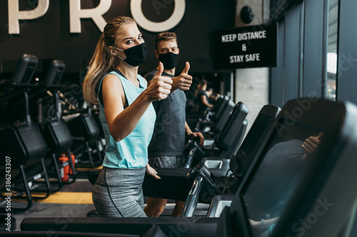 Young fit woman and man running on treadmill in modern fitness gym. They keeping distance and wearing protective face masks. Coronavirus world pandemic and sport theme. photo