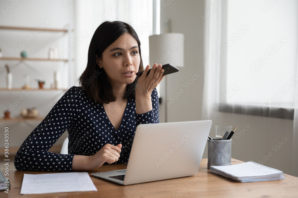 Confident young asian woman work on computer at home office make call talk on loudspeaker with colleague or client. Millennial Vietnamese female employee record voice audio message on smartphone. Confident young asian woman work on computer at home office make call talk on loudspeaker with colleague or client. Millennial Vietnamese female employee record voice audio message on smartphone.
