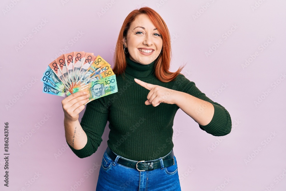 Beautiful redhead woman holding australian dollars smiling happy pointing with hand and finger Beautiful redhead woman holding australian dollars smiling happy pointing with hand and finger