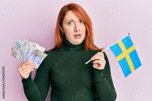 Beautiful redhead woman holding 20 swedish krona banknotes and sweden flag in shock face, looking skeptical and sarcastic, surprised with open mouth photo