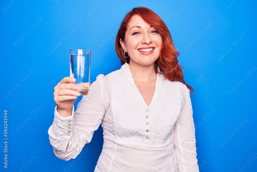 Young beautiful redhead woman drinking glass of water over isolated blue background looking positive and happy standing and smiling with a confident smile showing teeth Young beautiful redhead woman drinking glass of water over isolated blue background looking positive and happy standing and smiling with a confident smile showing teeth