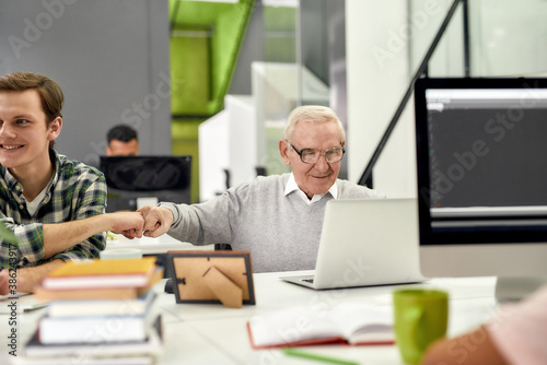 Aged man, senior intern looking at the screen of his laptop and doing a fist bump with his young colleague, Friendly male worker engaging new employee while working in the office