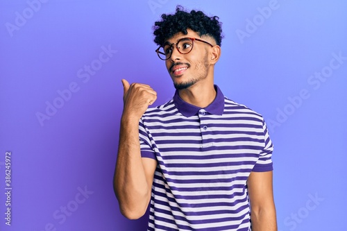 Young arab handsome man wearing casual clothes and glasses pointing thumb up to the side smiling happy with open mouth