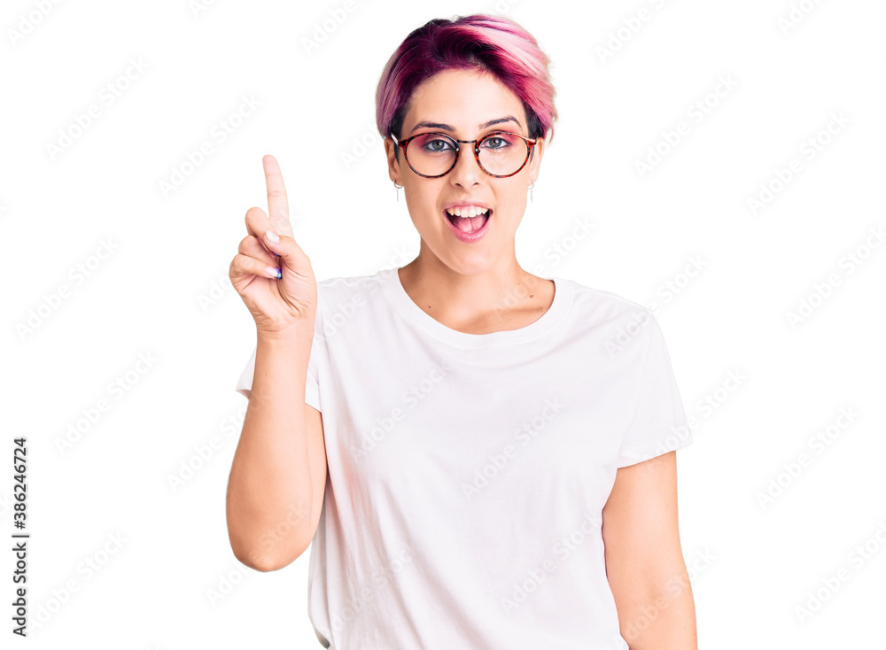 Young beautiful woman with pink hair wearing casual clothes and glasses showing and pointing up with finger number one while smiling confident and happy. Young beautiful woman with pink hair wearing casual clothes and glasses showing and pointing up with finger number one while smiling confident and happy.