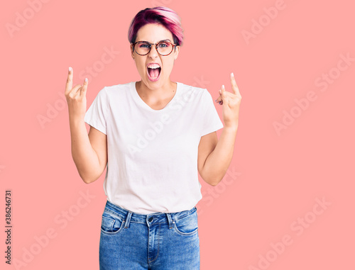 Young beautiful woman with pink hair wearing casual clothes and glasses shouting with crazy expression doing rock symbol with hands up. music star. heavy concept.