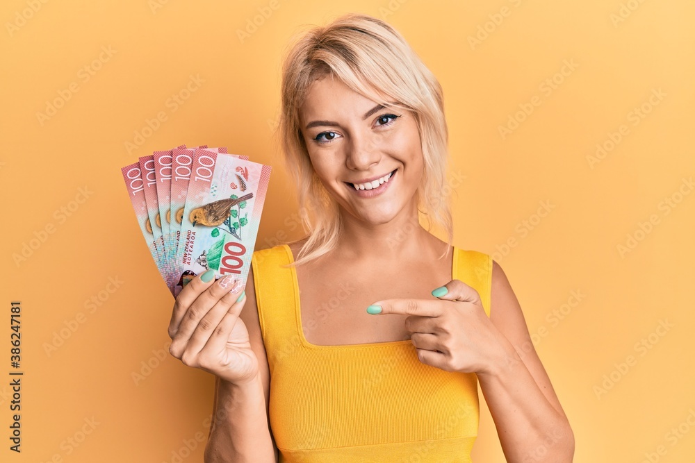 Young blonde girl holding 100 new zealand dollars banknote smiling happy pointing with hand and finger Young blonde girl holding 100 new zealand dollars banknote smiling happy pointing with hand and finger