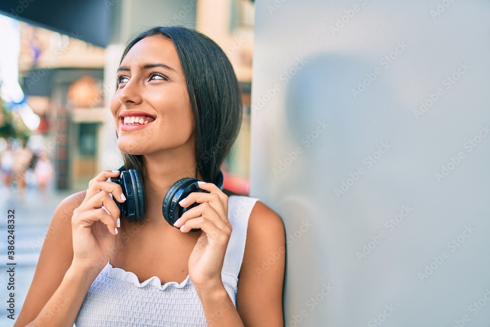 Young latin girl smiling happy using headphones leaning on the wall.