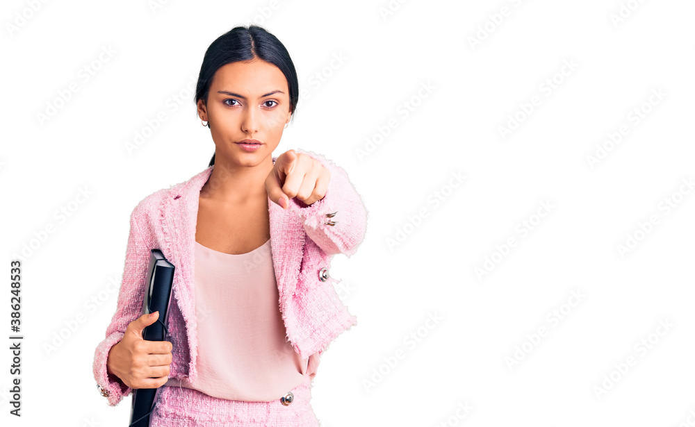 Young beautiful latin girl wearing business clothes holding binder pointing with finger to the camera and to you, confident gesture looking serious Young beautiful latin girl wearing business clothes holding binder pointing with finger to the camera and to you, confident gesture looking serious