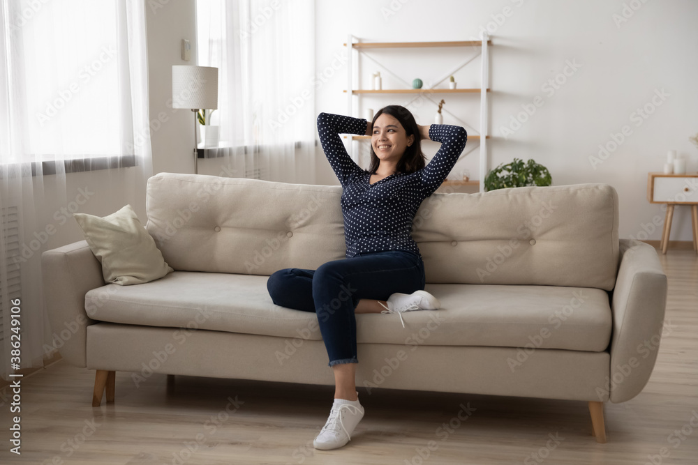 Happy millennial Vietnamese girl sit relax on sofa in living room look in window distance dreaming thinking. Smiling young asian woman renter rest on couch at home visualize imagine on weekend. Happy millennial Vietnamese girl sit relax on sofa in living room look in window distance dreaming thinking. Smiling young asian woman renter rest on couch at home visualize imagine on weekend.
