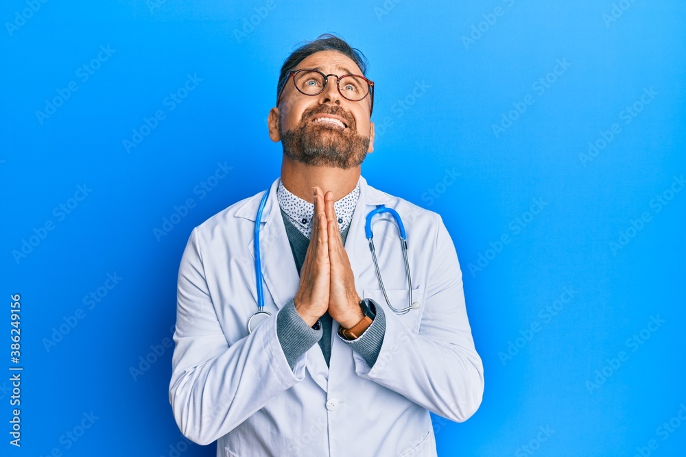Handsome middle age man wearing doctor uniform and stethoscope begging and praying with hands together with hope expression on face very emotional and worried. begging. Handsome middle age man wearing doctor uniform and stethoscope begging and praying with hands together with hope expression on face very emotional and worried. begging.