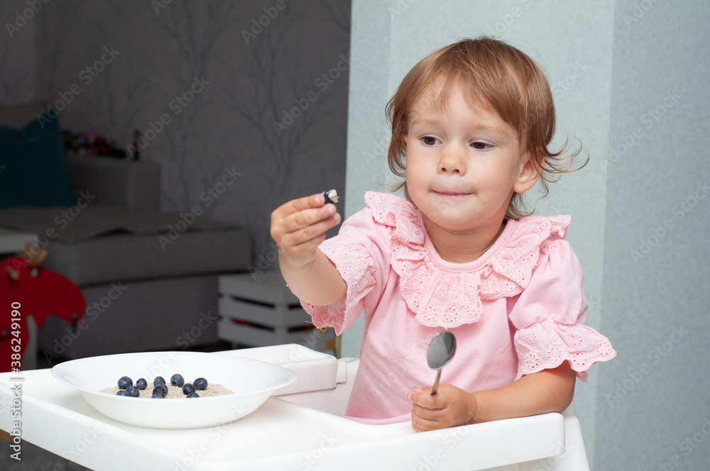 Happy, smiling child eating oatmeal with berries. The concept of a healthy breakfast for children, porridge with blueberries on baby chair Happy, smiling child eating oatmeal with berries. The concept of a healthy breakfast for children, porridge with blueberries on baby chair