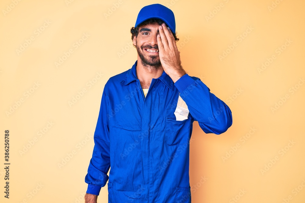 Handsome young man with curly hair and bear wearing builder jumpsuit uniform covering one eye with hand, confident smile on face and surprise emotion. Handsome young man with curly hair and bear wearing builder jumpsuit uniform covering one eye with hand, confident smile on face and surprise emotion.
