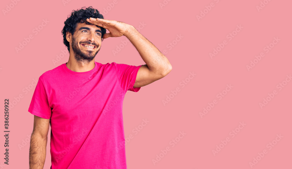 Handsome young man with curly hair and bear wearing casual pink tshirt very happy and smiling looking far away with hand over head. searching concept. Handsome young man with curly hair and bear wearing casual pink tshirt very happy and smiling looking far away with hand over head. searching concept.
