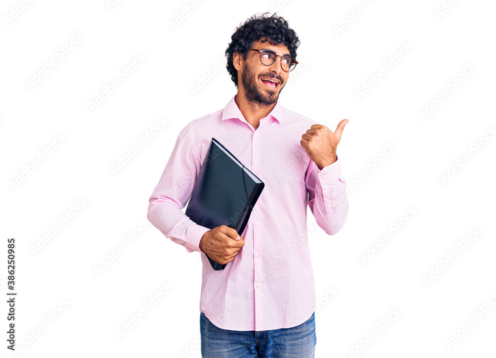 Handsome young man with curly hair and bear holding business folder pointing thumb up to the side smiling happy with open mouth Handsome young man with curly hair and bear holding business folder pointing thumb up to the side smiling happy with open mouth