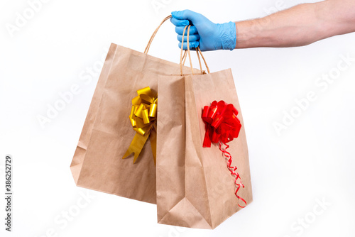 Secure contactless remote delivery of holiday gifts during the coronavirus pandemic. Close up. A courier hand in protective medical gloves holds two paper gift bag with a bow on a white background