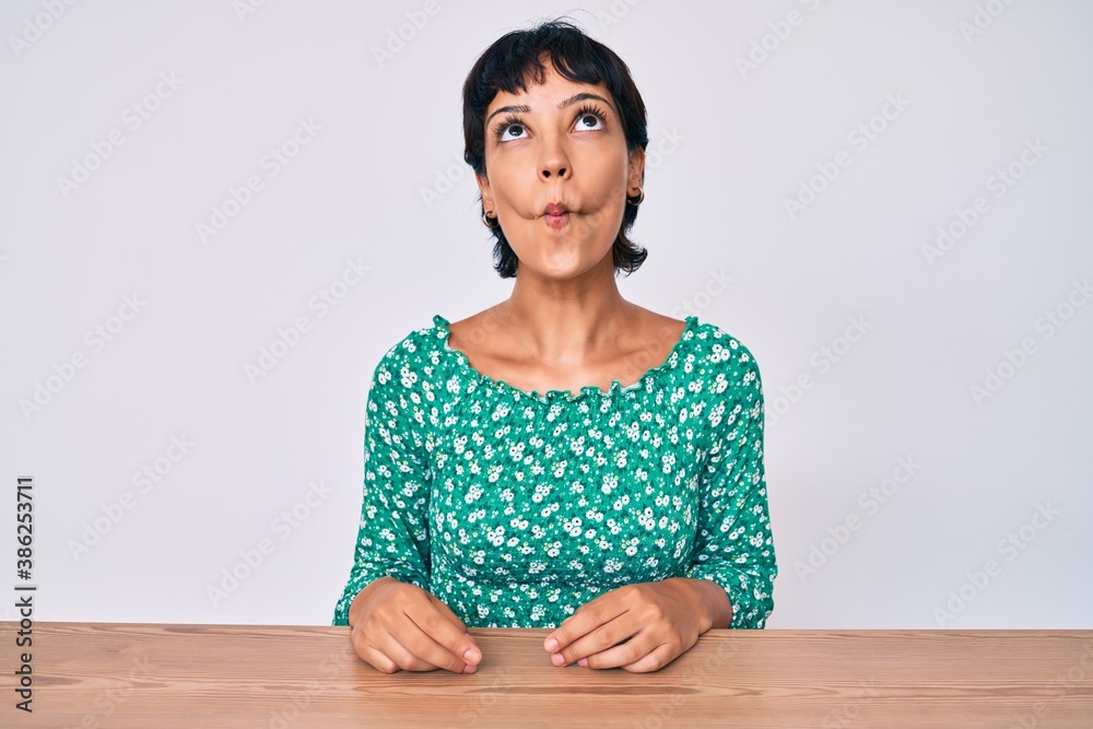 Beautiful brunettte woman wearing casual clothes sitting on the table making fish face with lips, crazy and comical gesture. funny expression. Beautiful brunettte woman wearing casual clothes sitting on the table making fish face with lips, crazy and comical gesture. funny expression.