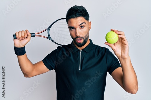 Young man with beard playing tennis holding racket and ball in shock face, looking skeptical and sarcastic, surprised with open mouth © Krakenimages.com