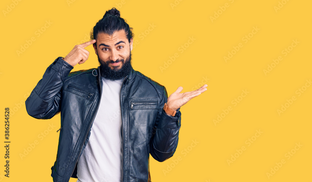 Young arab man wearing casual leather jacket confused and annoyed with open palm showing copy space and pointing finger to forehead. think about it.