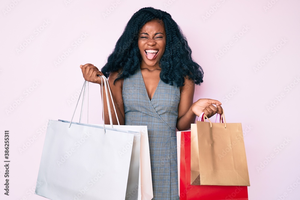 Young african american woman holding shopping bags sticking tongue out happy with funny expression. Young african american woman holding shopping bags sticking tongue out happy with funny expression.