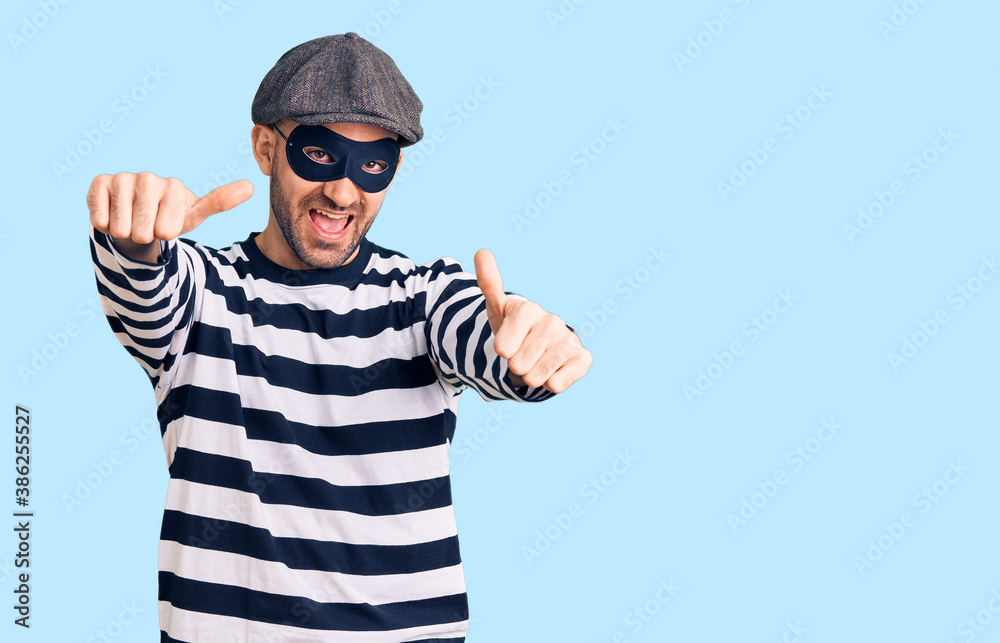 Young handsome man wearing burglar mask approving doing positive gesture with hand, thumbs up smiling and happy for success. winner gesture. Young handsome man wearing burglar mask approving doing positive gesture with hand, thumbs up smiling and happy for success. winner gesture.
