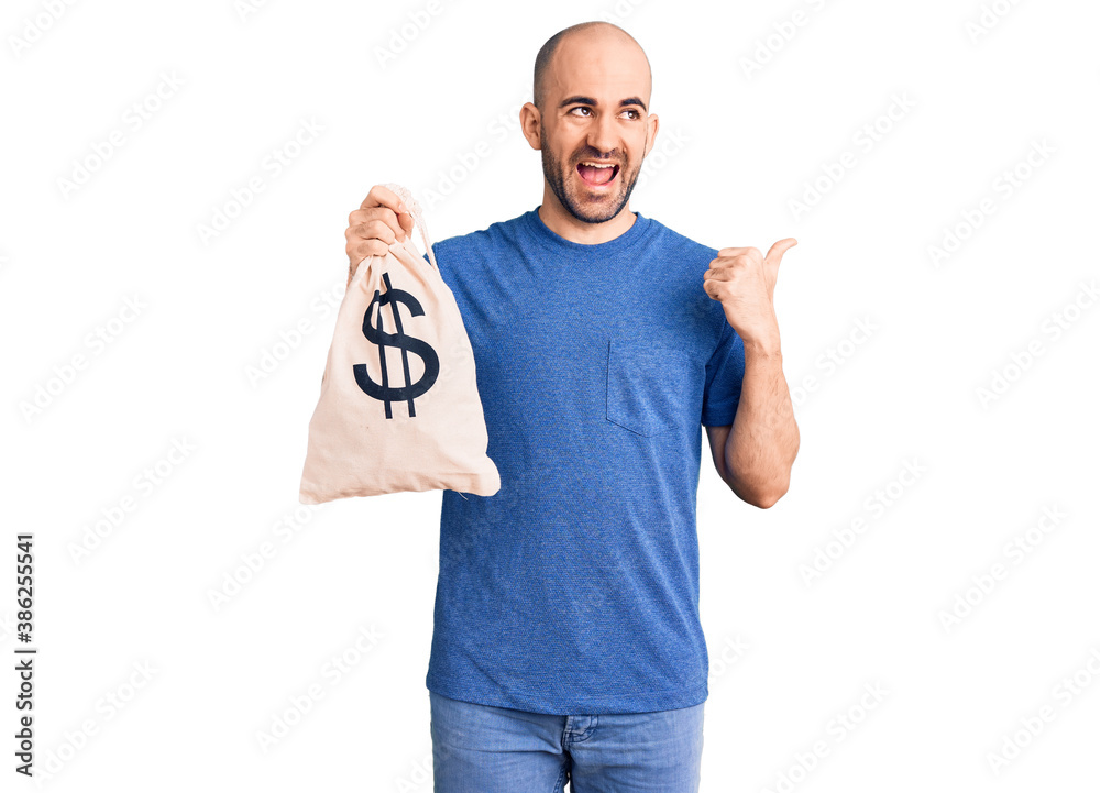 Young handsome man holding money bag with dollar symbol pointing thumb up to the side smiling happy with open mouth Young handsome man holding money bag with dollar symbol pointing thumb up to the side smiling happy with open mouth