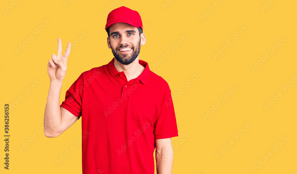 Young handsome man with beard wearing delivery uniform showing and pointing up with fingers number two while smiling confident and happy.