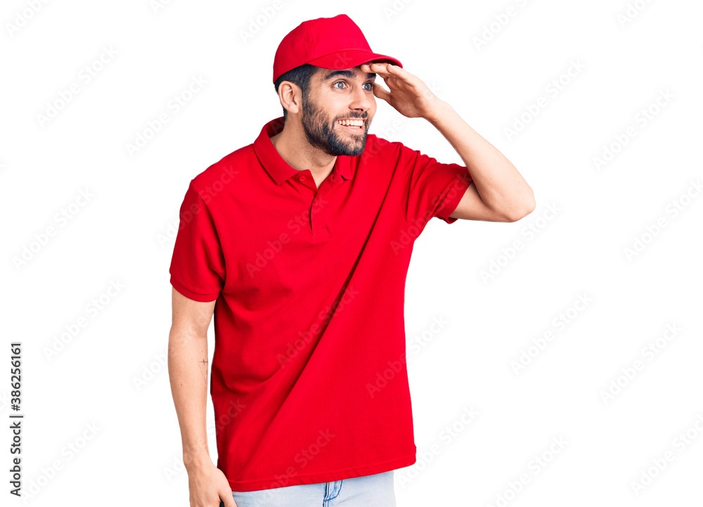 Young handsome man with beard wearing delivery uniform very happy and smiling looking far away with hand over head. searching concept.