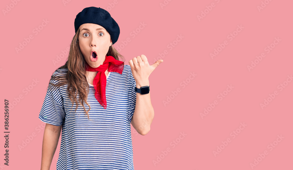 Young beautiful blonde woman wearing french beret and striped t-shirt surprised pointing with hand finger to the side, open mouth amazed expression. Young beautiful blonde woman wearing french beret and striped t-shirt surprised pointing with hand finger to the side, open mouth amazed expression.