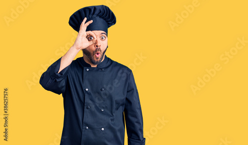 Young hispanic man wearing cooker uniform doing ok gesture shocked with surprised face, eye looking through fingers. unbelieving expression.