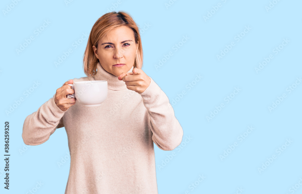 Young blonde woman holding coffee pointing with finger to the camera and to you, confident gesture looking serious Young blonde woman holding coffee pointing with finger to the camera and to you, confident gesture looking serious