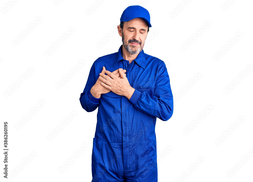 Middle age handsome man wearing mechanic uniform smiling with hands on chest with closed eyes and grateful gesture on face. health concept.