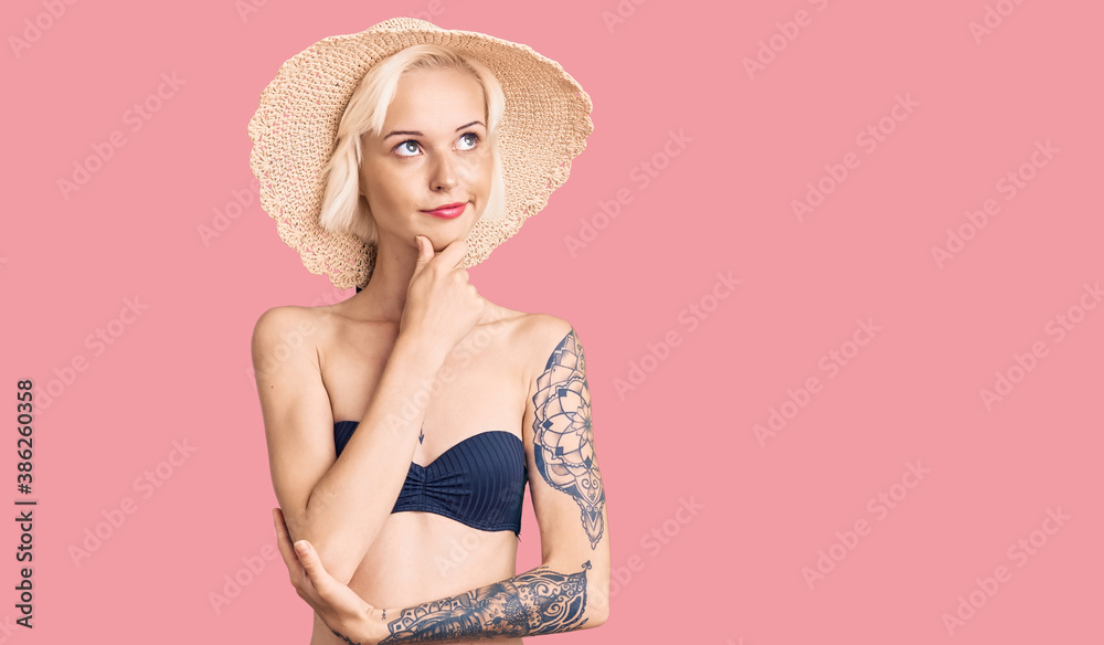 Young blonde woman with tattoo wearing bikini and summer hat with hand on chin thinking about question, pensive expression. smiling with thoughtful face. doubt concept. Young blonde woman with tattoo wearing bikini and summer hat with hand on chin thinking about question, pensive expression. smiling with thoughtful face. doubt concept.